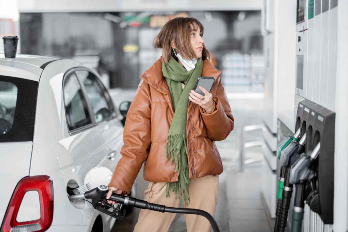 Eine junge Frau in einem braunen Ledermantel h&auml;lt die Zapfpistole in der rechten Hand, setzt sie in den Tankstutzen ihres Fahrzeugs ein, um aufzutanken, und h&auml;lt in der linken Hand ihr Handy, um den Benzinpreis in Island zu berechnen.