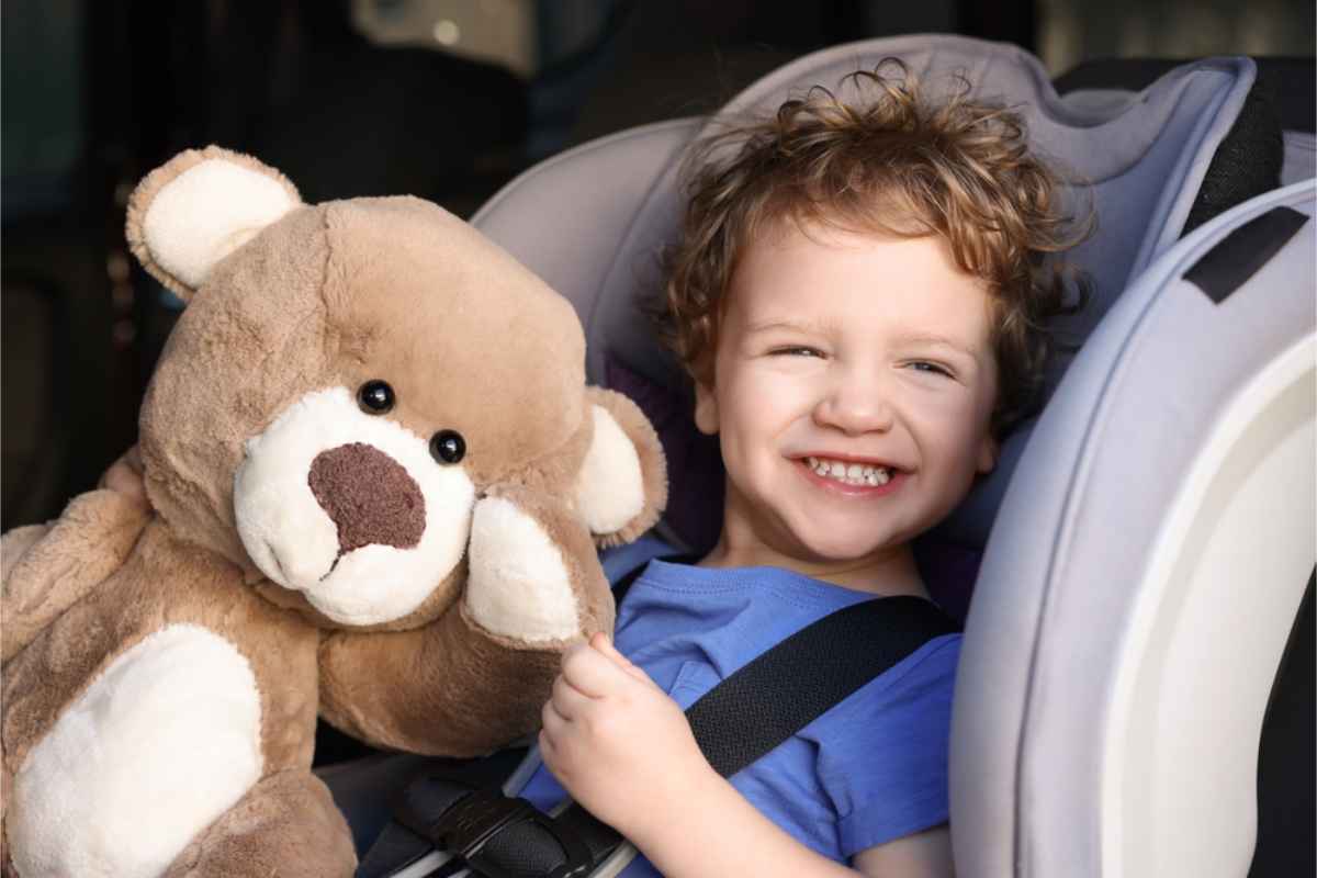 Glückliches Kind sitzt in seinem Kindersitz zusammen mit einem Teddybären